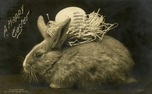 A Happy Easter card from the Osborne Collection of Early Children's Literature featuring a fluffy bunny with a chicken's egg in a nest of straw on its back, behind its little ears.