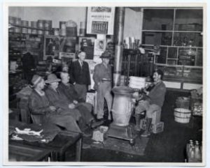 A man playing the fiddle in a country store in Tennessee in the 1940s. He sits next to a stove, a number of people sit or lean while giving him their undivided attention.