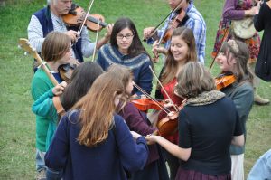 Shows a group of young teens delightedly playing the fiddle outdoors, on the grass. In the background, some adults play as well.
